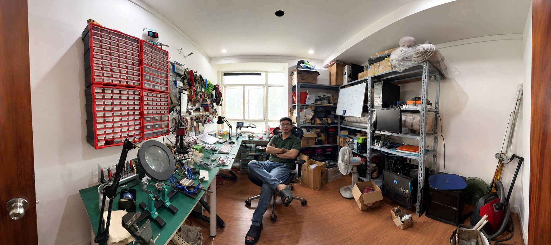 The SenseTechLab workshop. A wall of red and grey parts drawers above a bench carrying a microscope, an illuminated magnifier and soldering equipment; metal shelving of boxed components, monitors and test gear along the far wall; and the founder seated at the centre of the room.